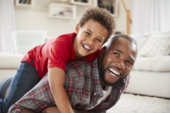 Son Climbs On Fathers Back As They Play Game In Lounge Together