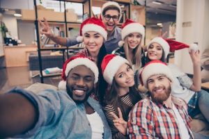 Self portrait of mixed race friends : african, american, asian, caucasian, young smiling bearded men and beautiful women in red christmas hat showing peace symbol to the camera, celebrating new year in the office